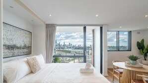 A modern hotel room with a large bed, a view of the cityscape, a dining area with a table and chairs, and a potted plant. - KozyGuru |  Rozelle | Bridge View with Carpark (Rozelle)