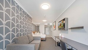 A hotel room with a grey patterned wall, a grey sofa, a bed, a desk, and a wall-mounted TV.