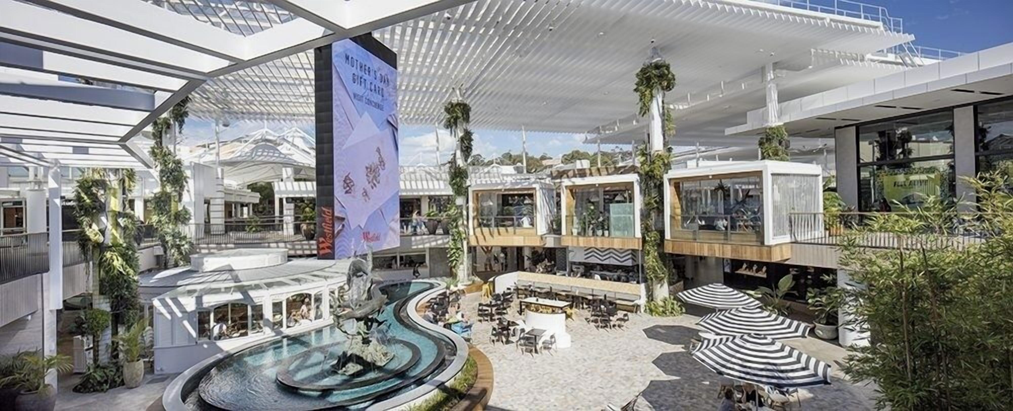 A modern hotel atrium with a central water feature, outdoor seating areas, and a Westfield shopping mall.