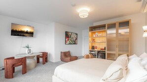 Quarto de hotel com cama, secretária com cadeiras, estante e televisão na parede.
