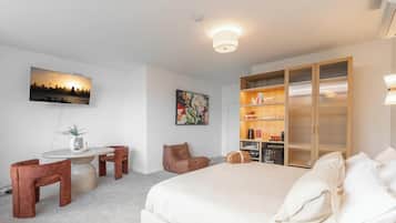 A hotel room with a bed, a desk with chairs, a bookshelf, and a TV on the wall.