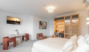 Habitación de hotel con cama, escritorio, sillas, estantería y televisión en la pared.