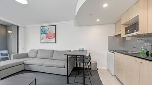A modern living room with a grey sofa, a small dining table, and a kitchen area with wooden cabinets and a sink.