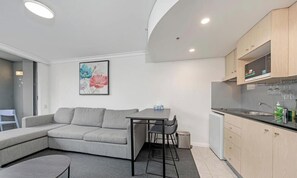 Una sala de estar moderna con un sofá gris, una pequeña mesa de comedor y una zona de cocina con armarios de madera y un fregadero.