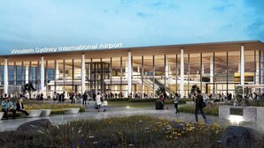 A modern airport terminal with a glass facade, a central staircase, and a landscaped area with seating and flowers.