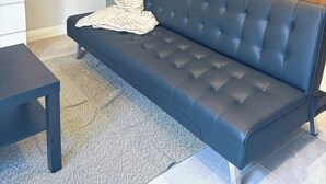A modern living room with a dark blue sofa, a small table, and a kitchen area with wooden cabinets and stainless steel appliances.
