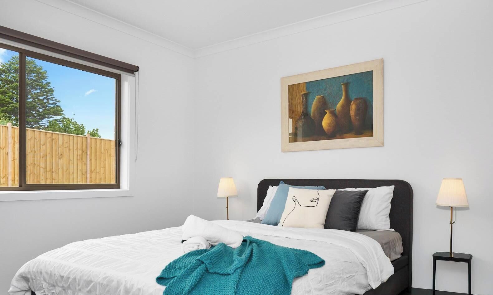 Ein Schlafzimmer mit einem Bett, zwei Nachttischlampen, einem Gemälde an der Wand und einem Fenster mit Blick auf Bäume.