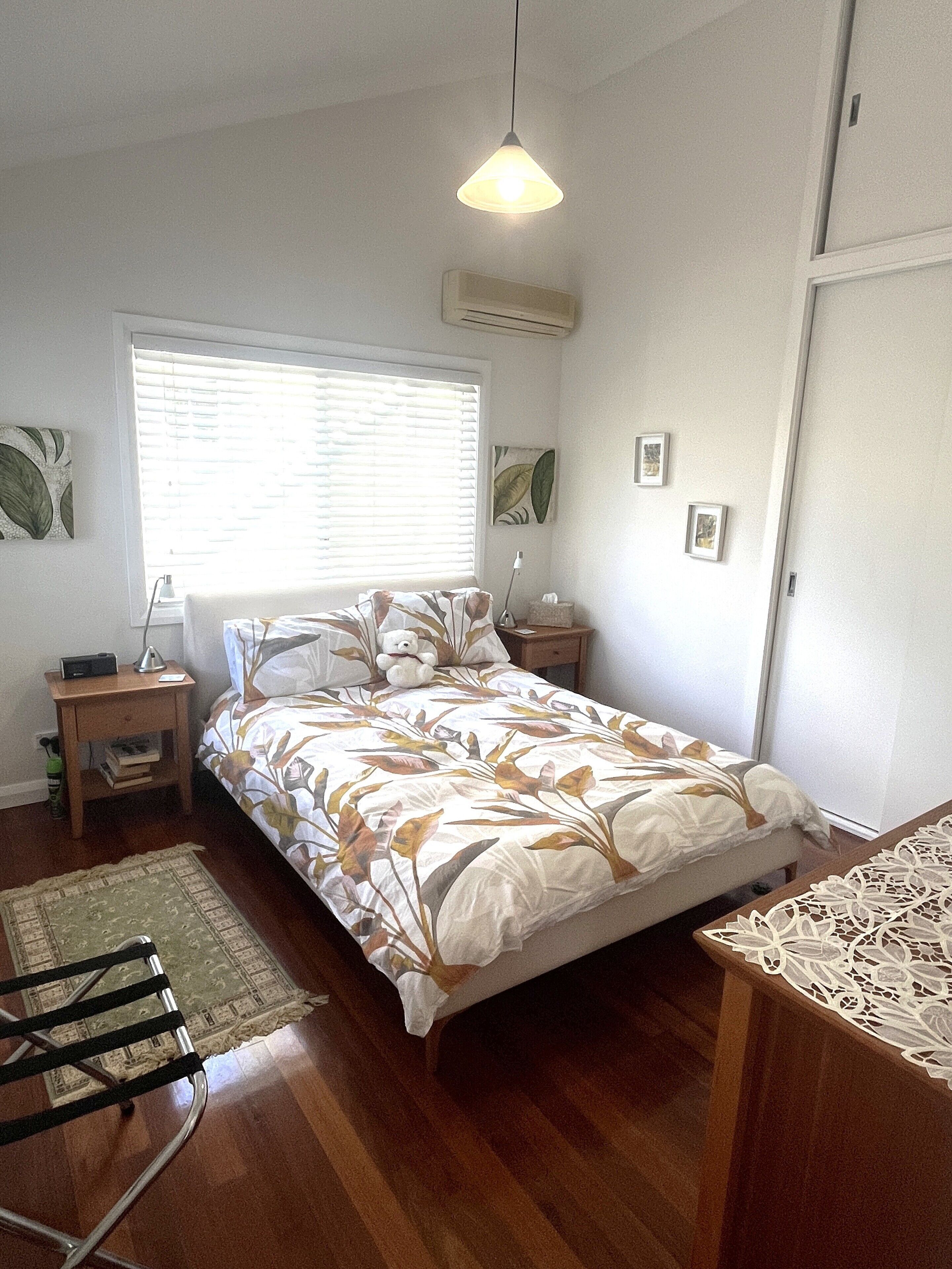 A bedroom with a bed, a bedside table, a lamp, a window with blinds, a wall-mounted air conditioner, and a door.
