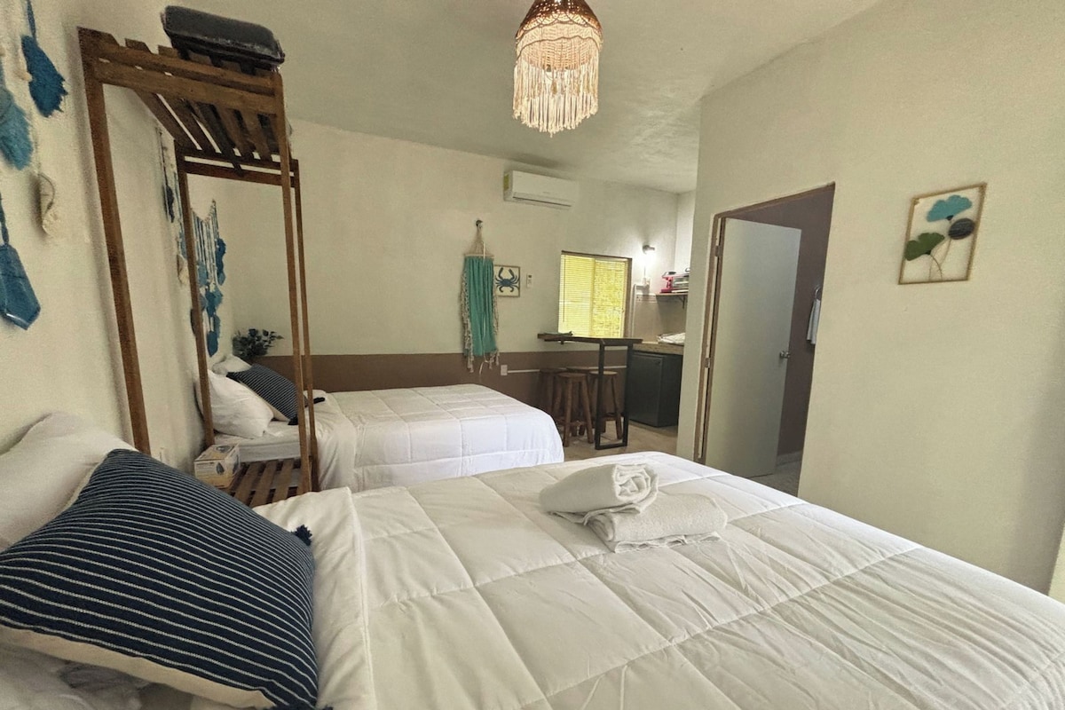 A room with two beds, a wooden ladder, a hanging light fixture, and a kitchenette in the background.