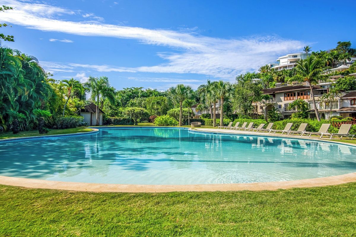 Une piscine entourée d’une végétation luxuriante, sous un ciel bleu et dégagé.