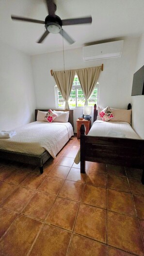 Quarto com duas camas, ventilador no teto, janela com cortinas e piso de azulejos.