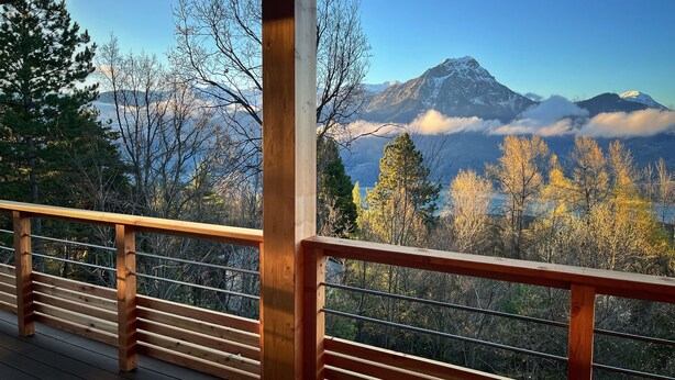 Eine Holzterrasse mit Geländer, die auf eine gebirgige Landschaft mit Bäumen und einem markanten Gipfel blickt.