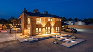 A modern wooden house with a garage, illuminated windows, and a car parked outside. - Cedar House | Pickleball, Saltwater Pool, Sauna (Paso Robles)