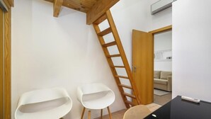 Une pièce avec une échelle en bois, deux chaises blanches, une table noire et, au fond, un canapé beige.