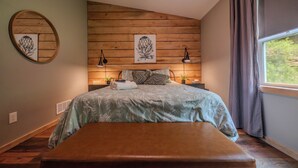 Een slaapkamer met een bed, een bankje, houten lambrisering aan de muren, twee wandlampen en een raam met gordijnen.