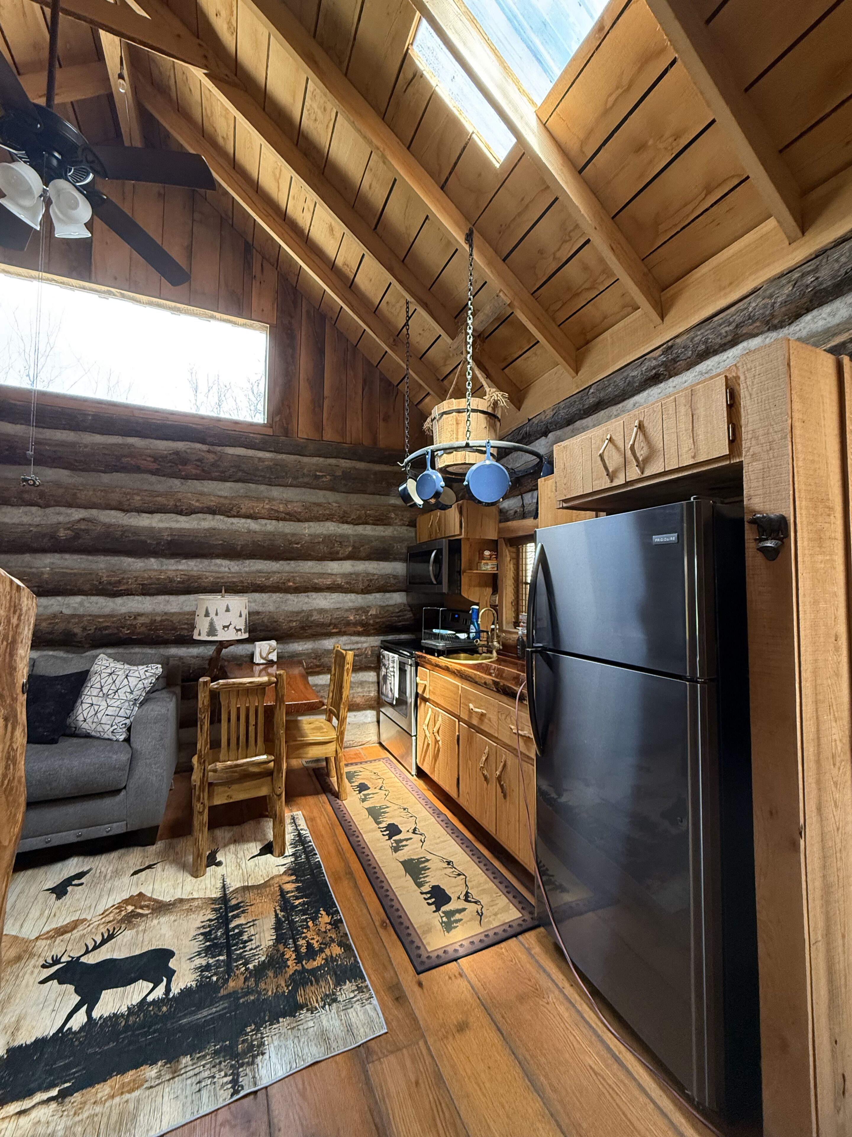 A log cabin interior with a wooden dining table, a sofa, a kitchen with a black refrigerator, and a hanging pot rack.