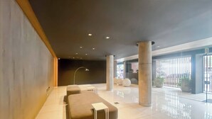 Eine moderne Lobby mit minimalistischem Design, ausgestattet mit einer Sitzgruppe, einer Bank und einem kleinen Tisch, großen Säulen und einer Glastrennwand mit Blick ins Freie.