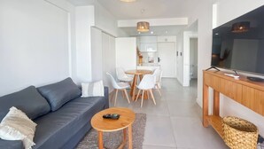 Ein modernes Wohnzimmer mit einem grauen Sofa, einem kleinen Holztisch, einem Fernseher auf einem Holzschrank und einem Essbereich mit einem weißen Tisch und Stühlen.