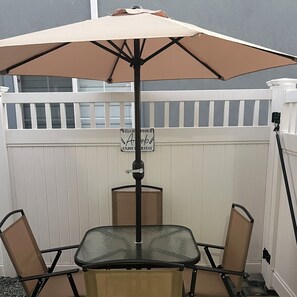 En uteplats med ett bord och stolar under ett stort parasoll, en skylt med texten ”Välkommen till Airbnb, hoppas ni får en trevlig vistelse” och ett vitt trästaket i bakgrunden.