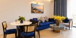 A living room with a round table, blue sofa, and chairs, a painting on the wall, and a window with curtains.