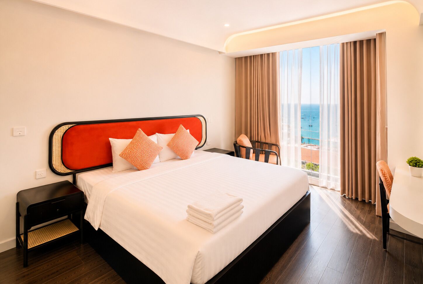 A hotel room with a bed, a nightstand, a chair, a small table with a plant, and a view of the sea.