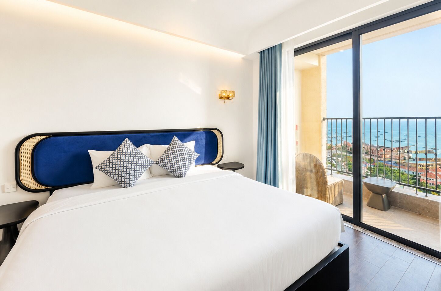 A hotel room with a large bed, a nightstand, a wall-mounted lamp, and a view of the sea through a balcony.