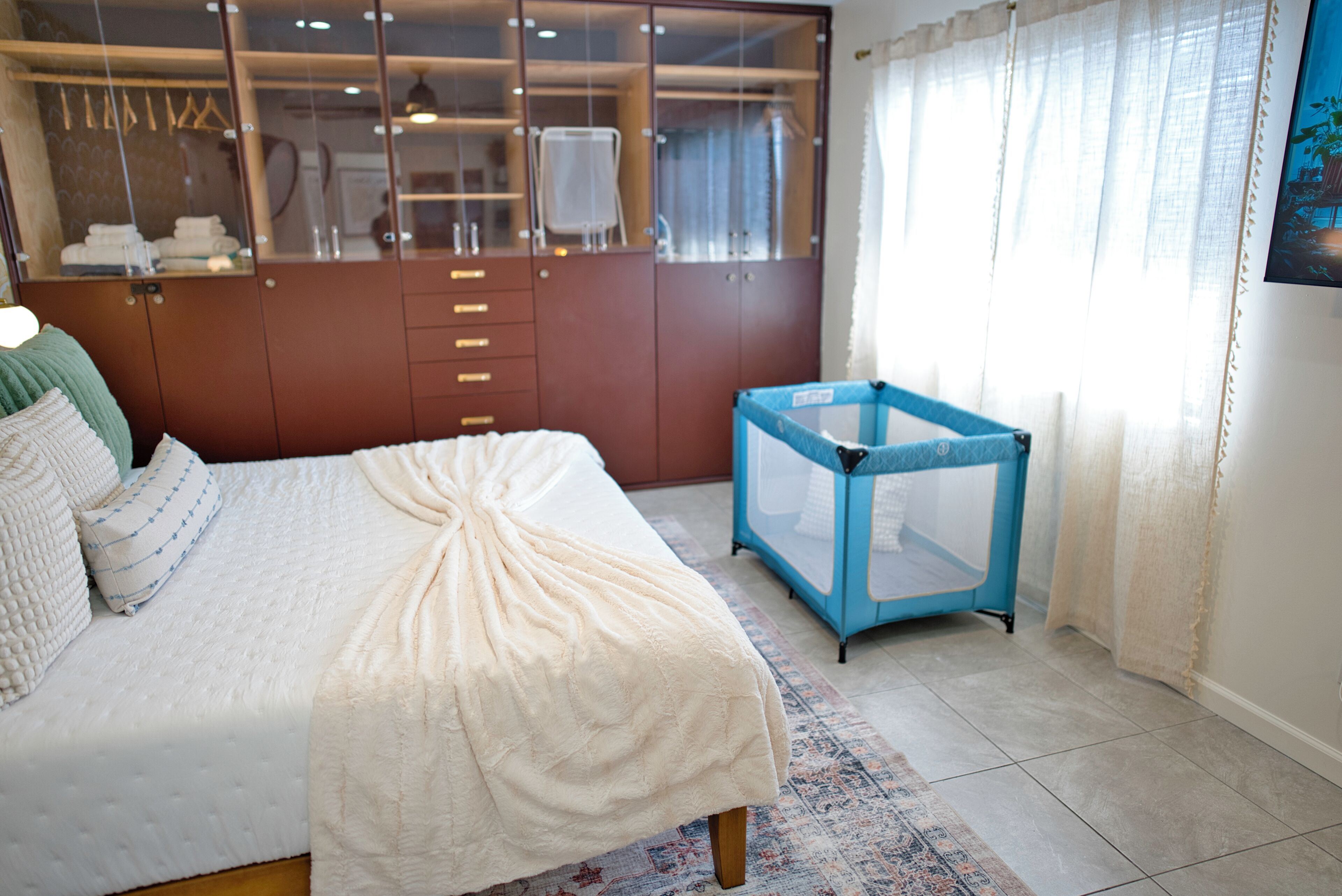 A bedroom with a bed, a wardrobe, a crib, and a window with curtains.