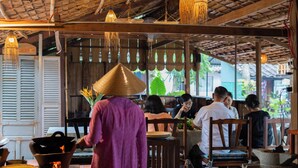A restaurant with wooden furniture, hanging lights, and a person cooking. - Bình An Village (Phuoc Hau)