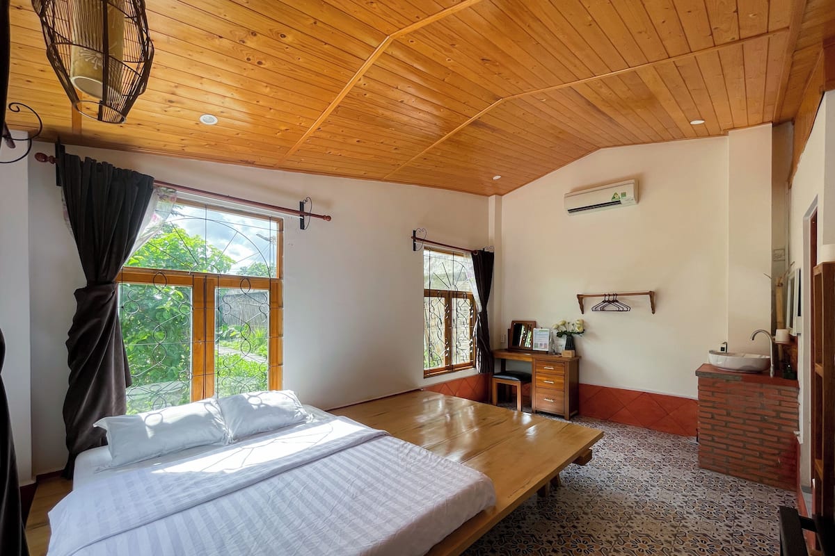 A bedroom with a bed, a desk, a chair, a window with curtains, and a bathroom with a sink and mirror.