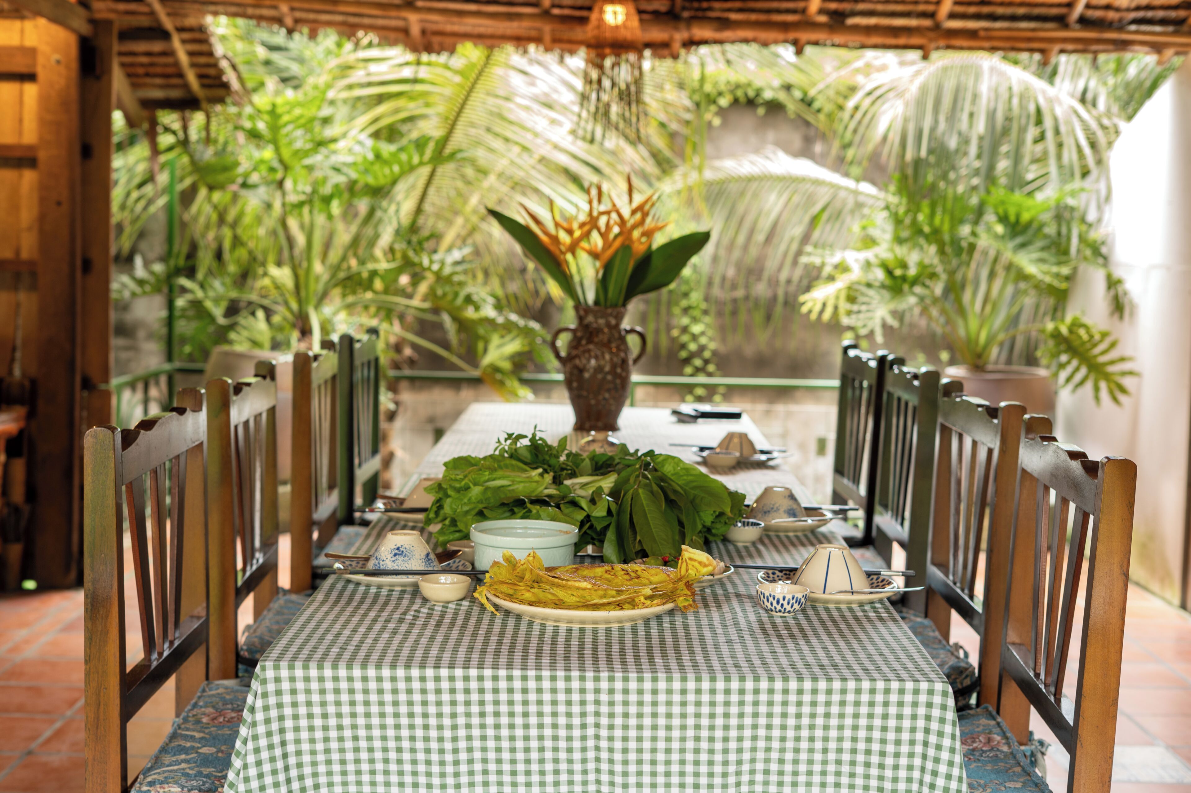 An outdoor dining area with a checkered tablecloth, wooden chairs, a vase with foliage, and a table set with plates and utensils.