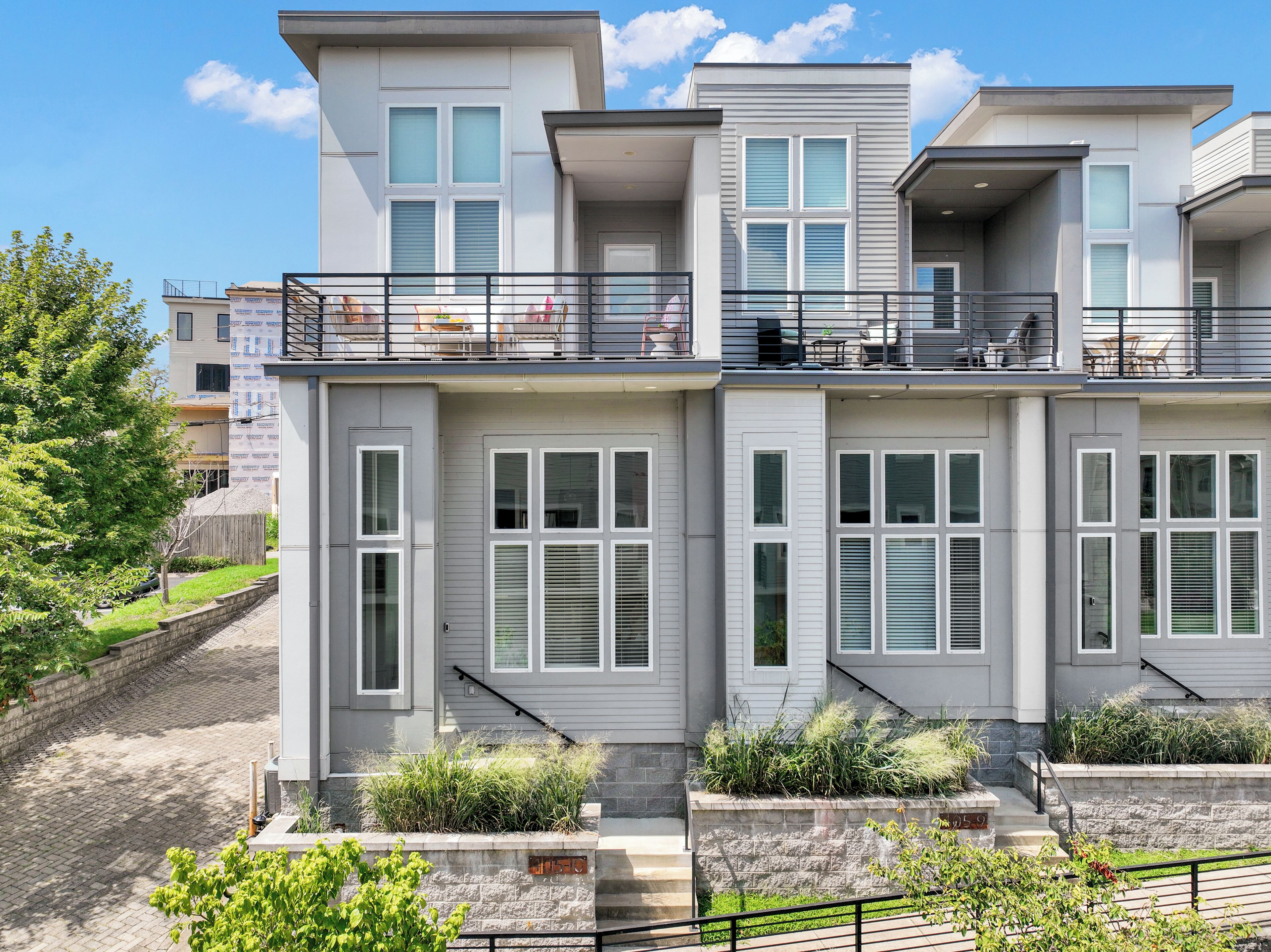 A modern multi-story residential building with a balcony, large windows, and a stone pathway leading to the entrance.