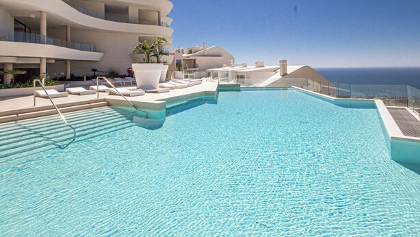 A modern infinity pool with clear blue water, surrounded by white lounge chairs and a white building with balconies.