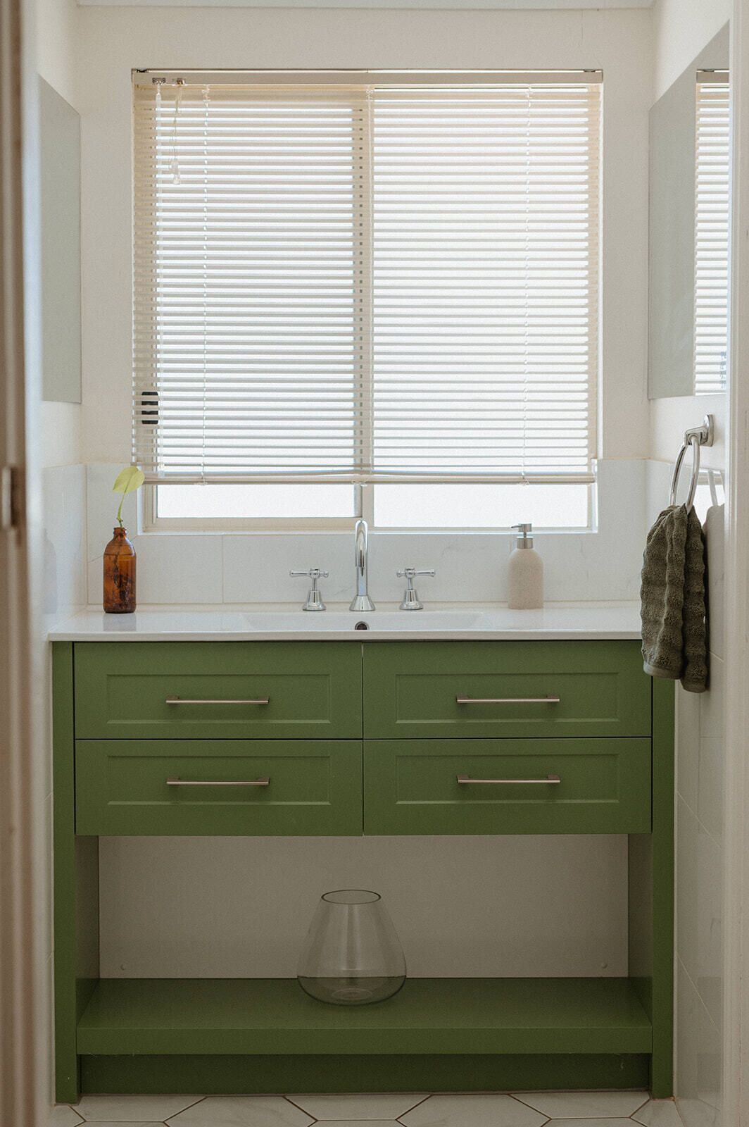 Une salle de bain avec un lavabo blanc, des meubles verts et une fenêtre avec des stores.
