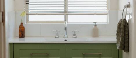 Une salle de bain avec un lavabo blanc, des meubles verts et une fenêtre avec des stores.