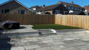 Un jardin avec une clôture en bois, une pelouse verdoyante et une terrasse dallée.
