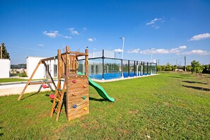 A playground with a wooden climbing structure and a slide, surrounded by a fenced area with a pool in the background.