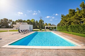 Una piscina rectangular con borde de azulejos, rodeada de sillas de descanso y una pérgola con luces colgantes.