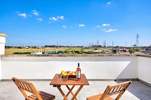 A rooftop terrace with a wooden table and chairs, a bottle, and a bowl of fruit.