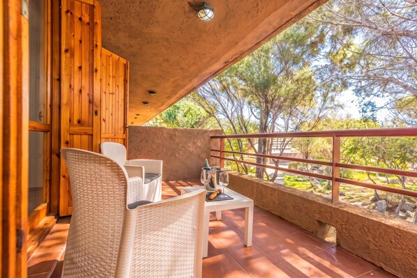 A balcony with wicker furniture, a table with a bottle and glasses, and a view of trees.