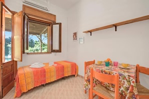 Een kamer met een bed, een tafel met een tafelkleed met een fruitmotief, en een raam met een open luik.