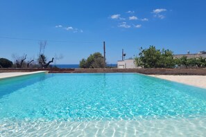A clear blue pool with a view of the sea, surrounded by trees and a building.