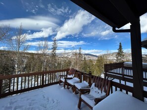 En snedækket terrasse med træstole og gelænder, med udsigt over et bjerglandskab med snedækkede træer og en klar blå himmel.