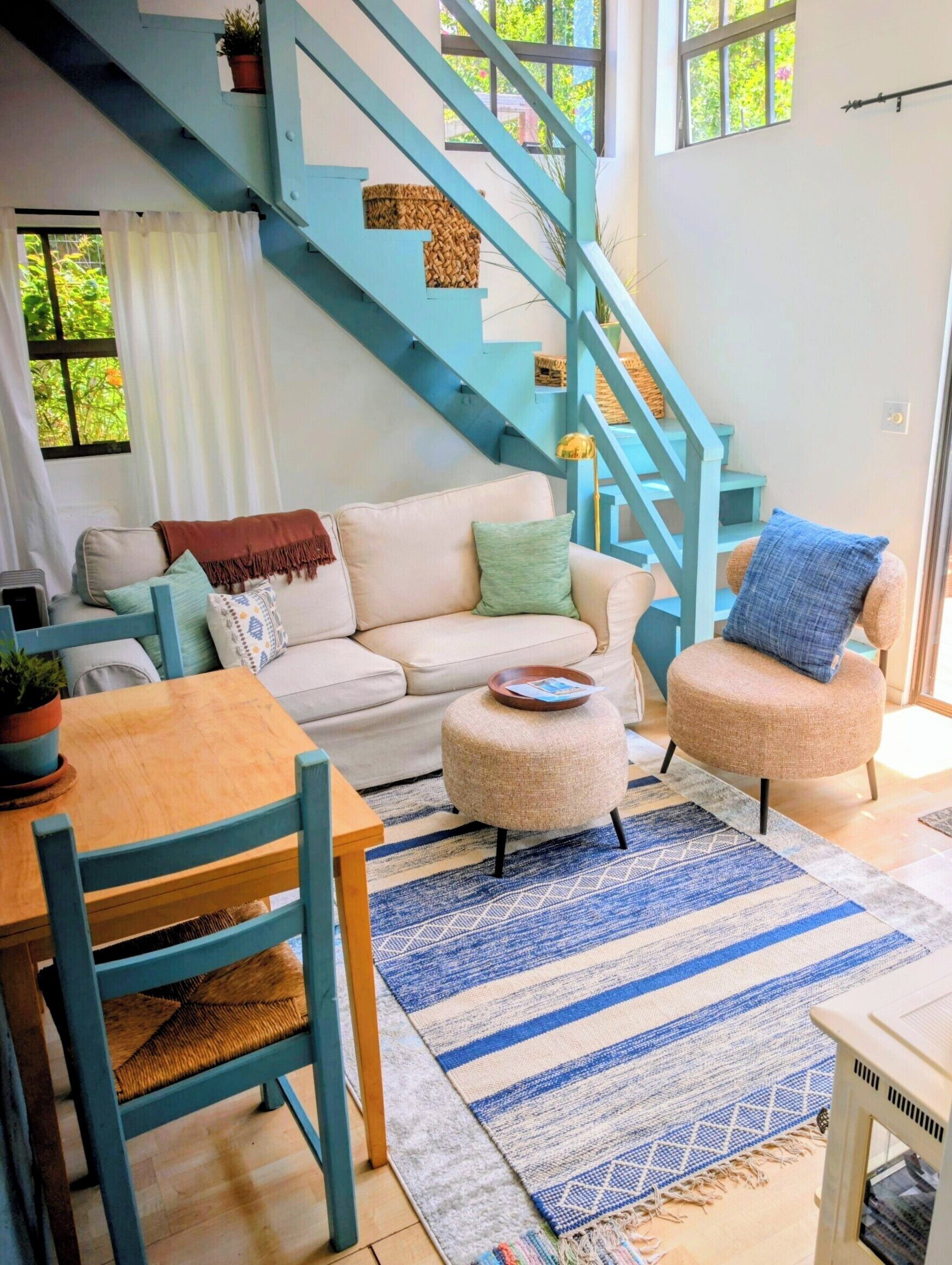Un salon avec un canapé, un pouf et une table en bois. On peut voir un escalier bleu avec un panier posé sur la plateforme.