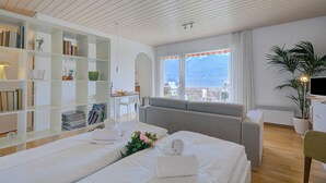 A hotel room with two beds, a sofa, a desk, a bookshelf, and a large window with a view. - Casa Miramonti Muralto with Garage and Garden, Muralto, Switzerland (Muralto)