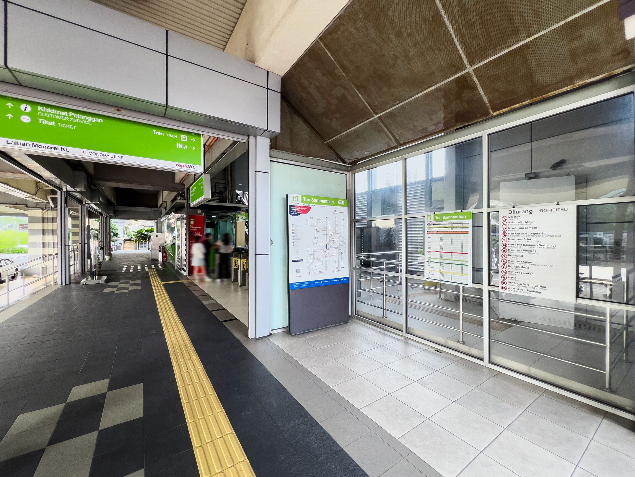 A train station platform with directional signs, a ticket counter, and a glass barrier.