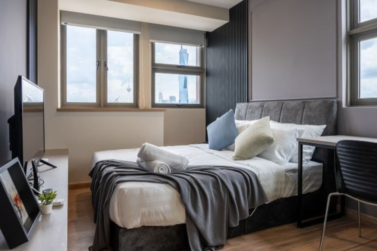 Un dormitorio moderno con cama, escritorio y televisor. Ventanas ofrecen vista a la ciudad.