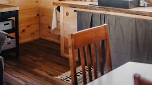 L’intérieur d’un chalet en bois comprend un coin cuisine avec une table en bois, des chaises, un porte-ustensiles suspendu et une fenêtre qui laisse entrer la lumière naturelle.