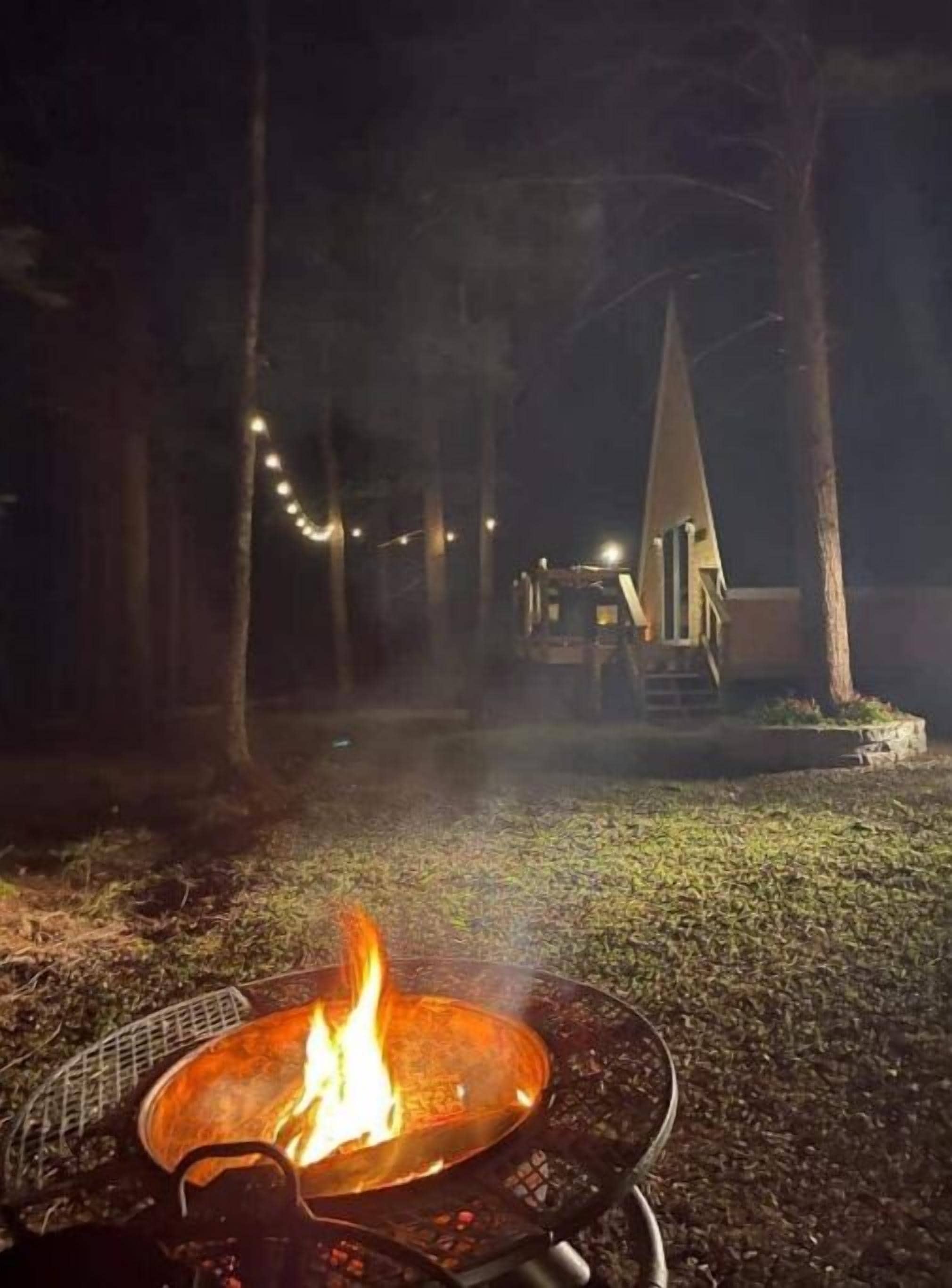 A campfire in a metal fire pit with a metal grill, surrounded by a grassy area and trees.