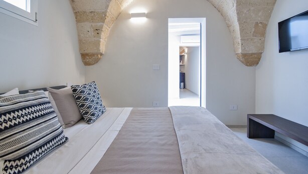 A bedroom with a stone arch ceiling, a bed with patterned pillows, a bench, and a TV on the wall.