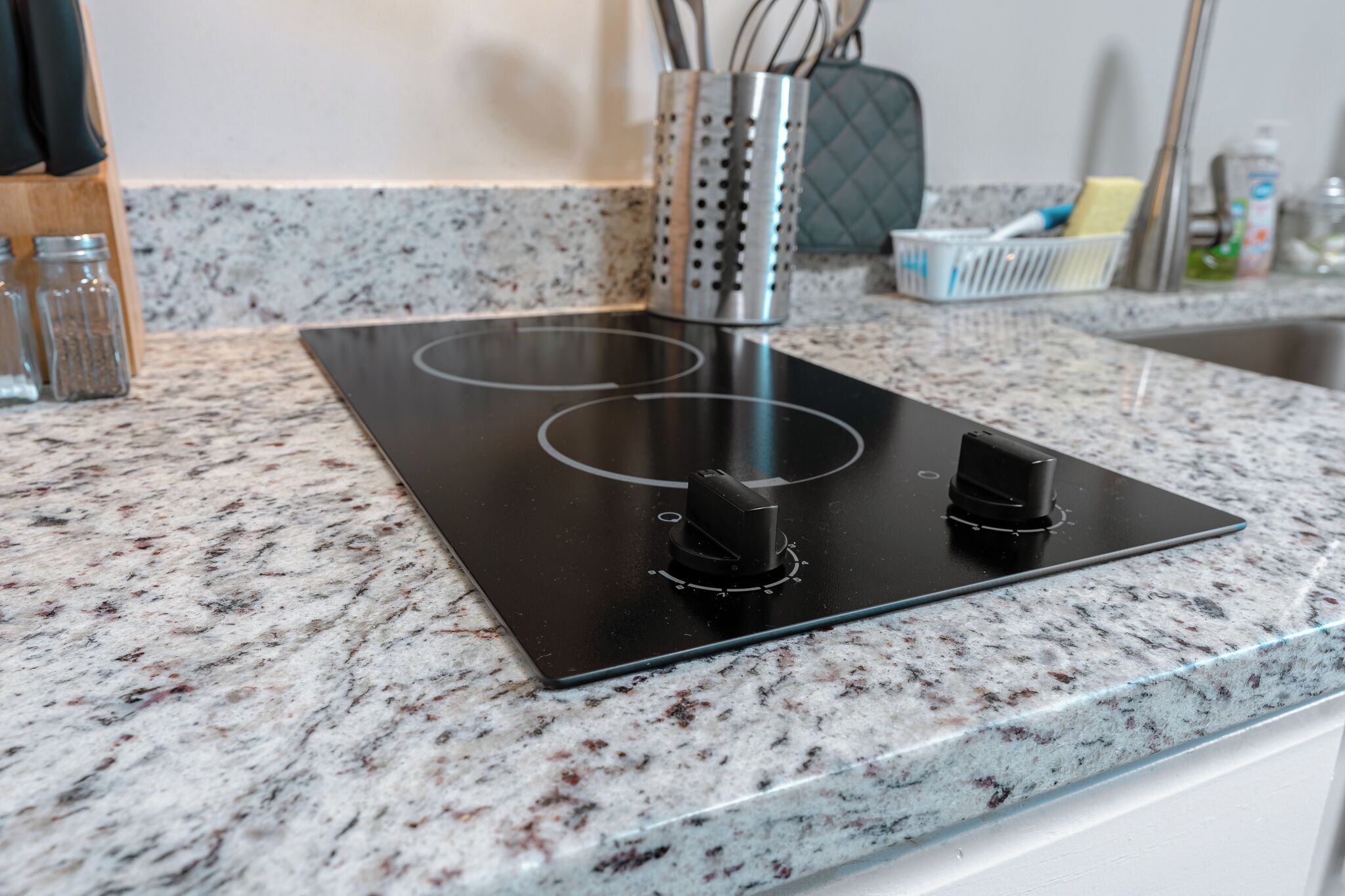 Une cuisine dotée d’une plaque de cuisson à induction noire, d’un plan de travail en granit et d’un porte-ustensiles en acier inoxydable.